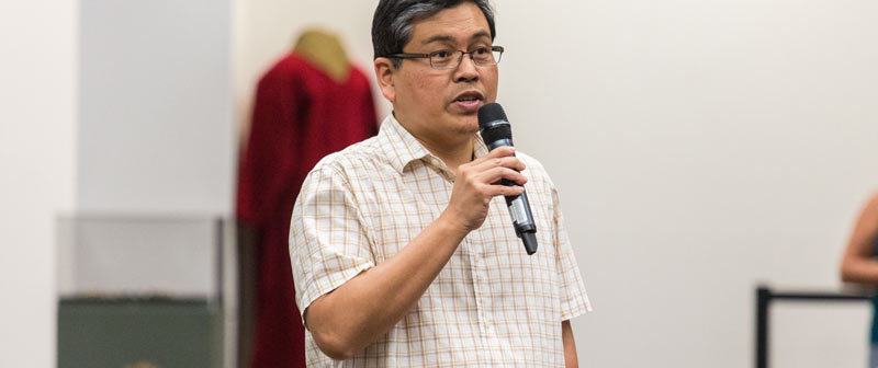 A man in a checked shirt and glasses speaks into a microphone while standing indoors.