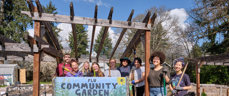 A group of people smiling and holding gardening tools stand together under a wooden pergola. In front of them is a sign that reads PLU Community Garden.