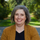 A person with short gray hair is smiling outdoors. They are wearing a mustard-colored cardigan over a black top. The background features a blurred path and greenery.