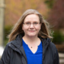 A person with long, light-brown hair and glasses is wearing a blue top and a black jacket. They are outdoors, with a blurred background of trees and path.