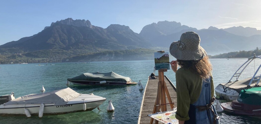 Student painting a hillscape on the water with boats using an easel.