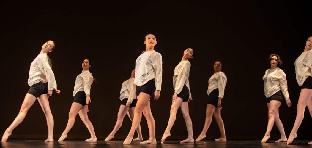 PLU's Dance team during their winter showcase. Each dancer takes a step forward, toes pointed. They are wearing white long-sleeved shirts and black shorts.