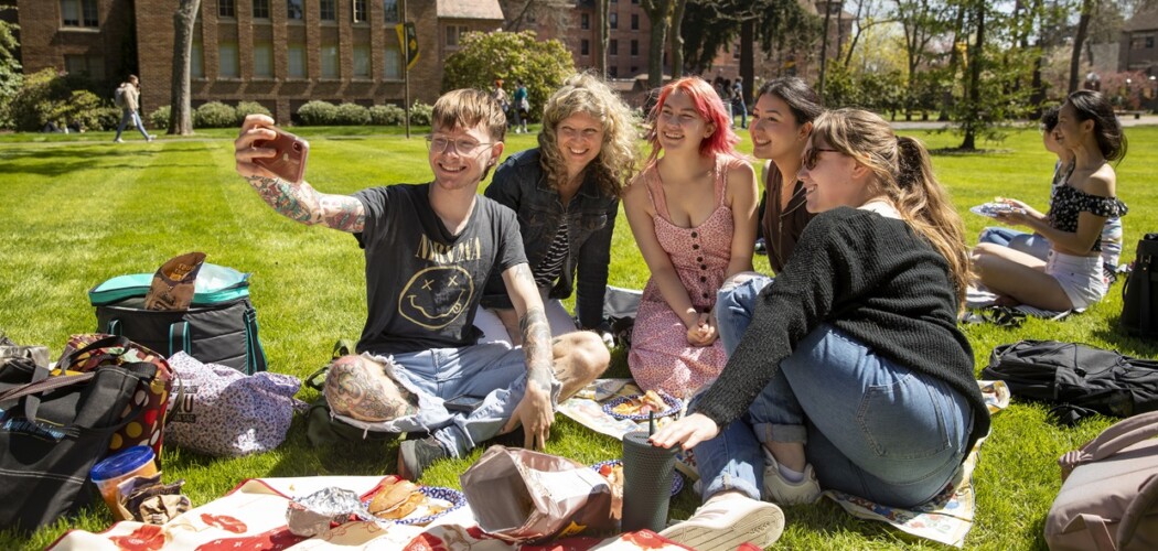 A group of French students along with their professor take a selfie outside on grass on campus during a French picnic for class