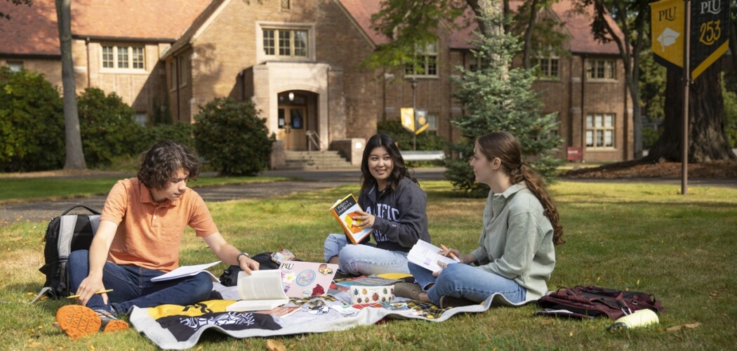 Three students sit outside on a blanket in the grass during a sunny day on campus. They have books open and seen to be studying or in conversation. Behind them is a brick academic building.