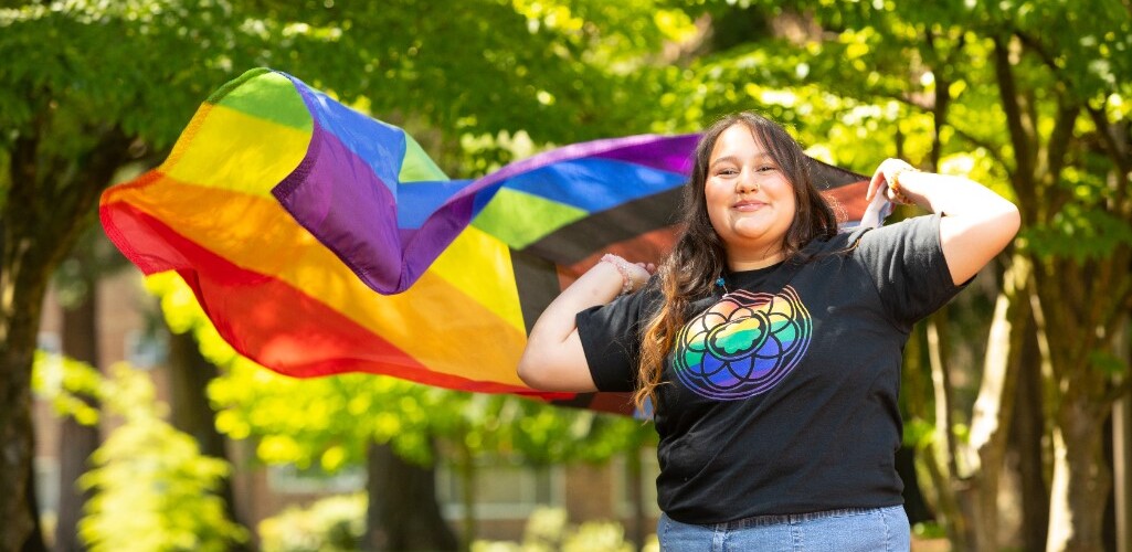 Student wearing PLU rainbow rose window shirt holding a pride flag behind their shoulders. The flag is seen by the wind blowing it to the side.