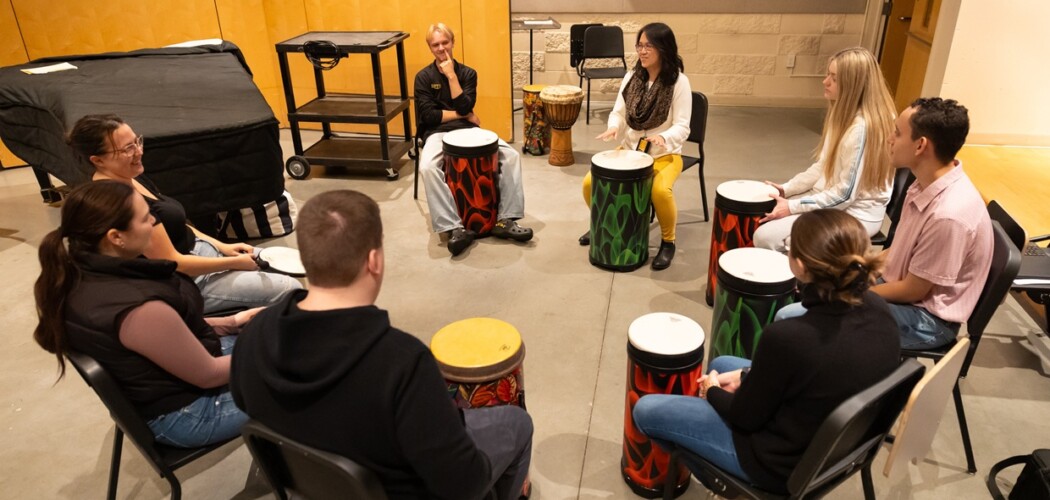 Students and a professor sit with drums in front of them in a circle during a 'Psychology of Music' class.