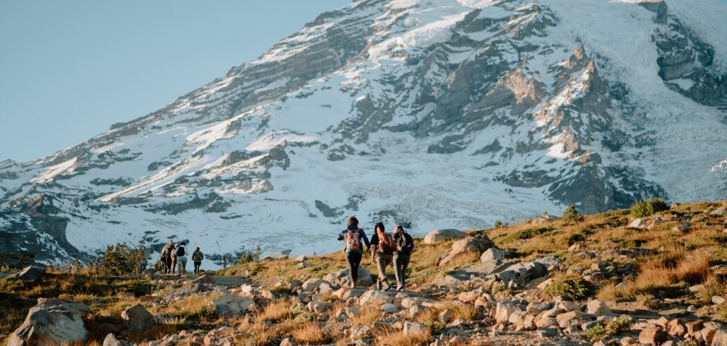 Students wearing backpacks walk along a rocky mountain trail. Ahead of them looms the large snow-capped peak of Mount Rainier