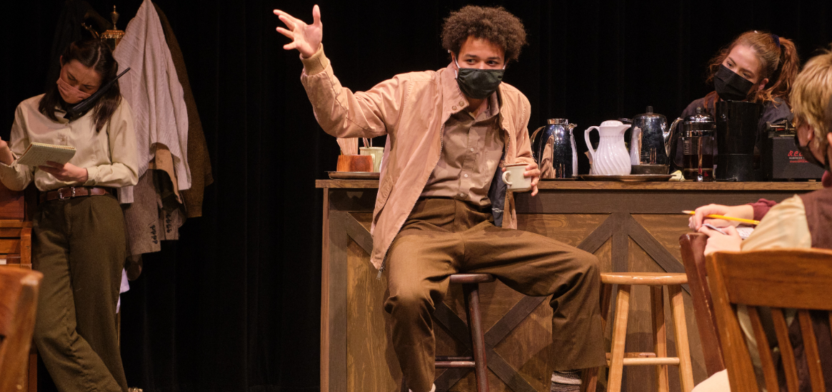 Student performing a monologue surrounded by ensemble within a cafe-style set in a performance of The Laramie Project in the Studio Theatre