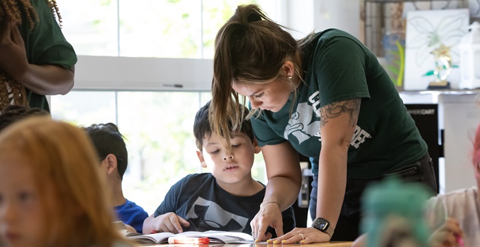 A student teacher stands at a desk with a young student. Both are looking at a book on the desk together. In the background are other students.
