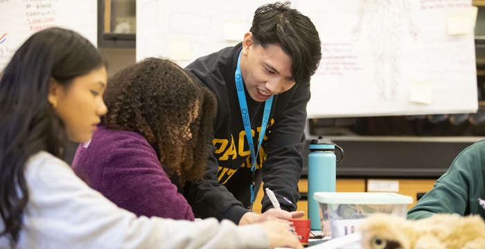 A PLU student teacher leans over a table with two middle school students.