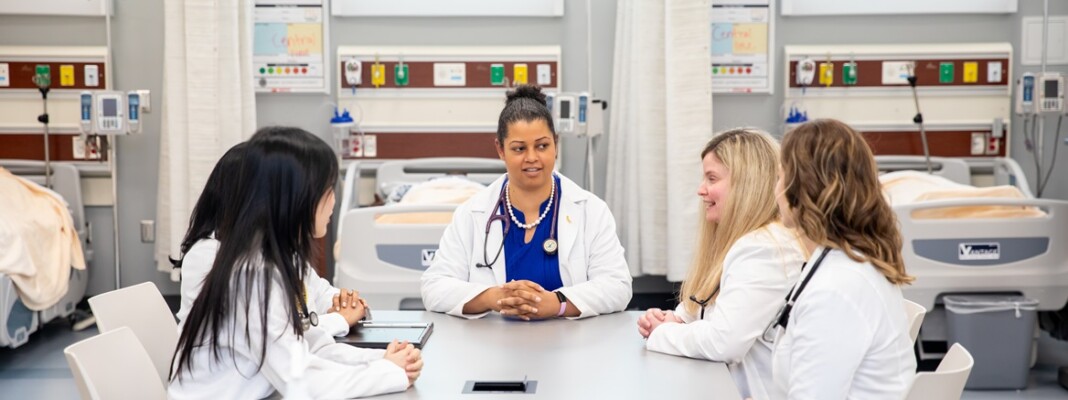 DNP students in white lab coats sit around a table and chat in PLU's Nursing Lab & Simulation Center