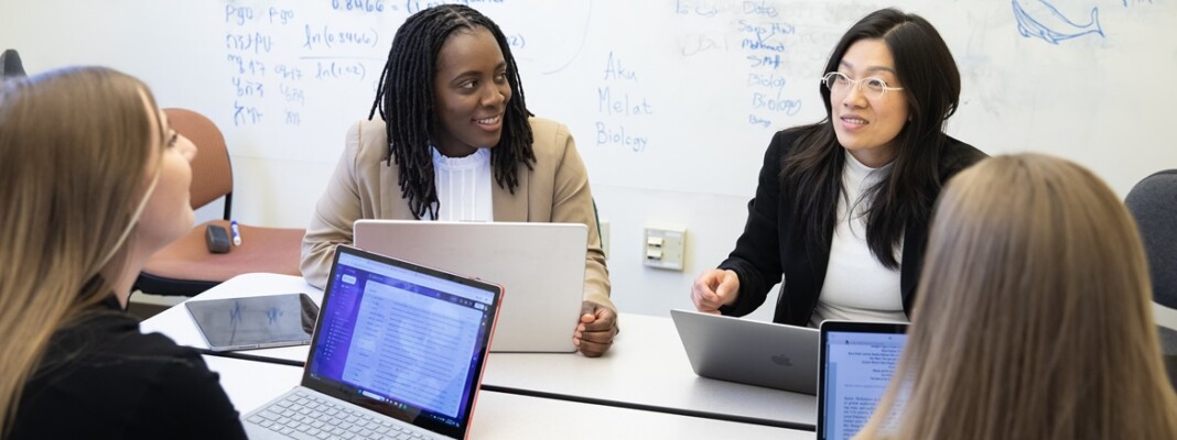 PLU MBA students dressed in business casual attire sit around a table and converse in a small classroom, laptops open in front of them