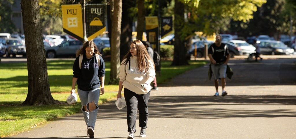 Two students wearing backpacks walk down a sidewalk on campus on a sunny day. In the background are black and yellow PLU banners on lightposts, as well as other students walking