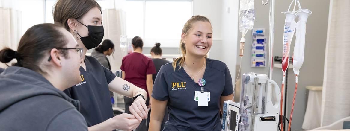 Three PLU nursing students stand near a bedside IV stand and drip bags in the PLU Nursing Simulation Center