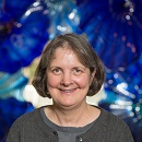 Cathy Krebs with shoulder-length hair smiles against a vibrant blue and purple abstract background. They are wearing a gray top.
