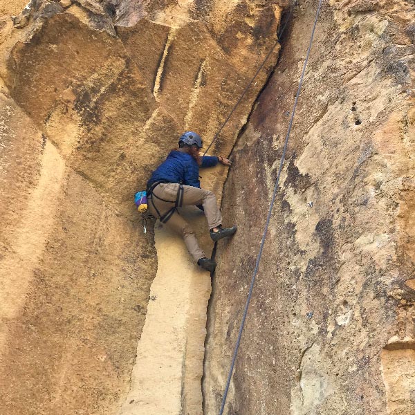 A person in a blue jacket and helmet climbs a rugged rock face, focused and determined. Climbing gear provides safety.