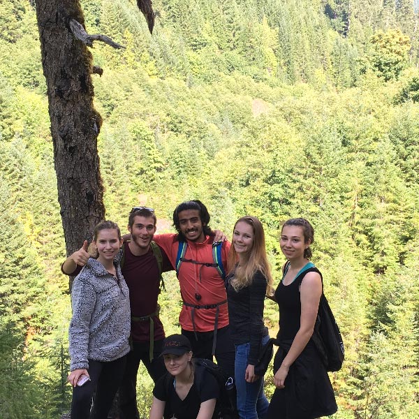 Five PLU students smiling on a hiking trip, standing together on a forest trail. Dense green trees cover the background.