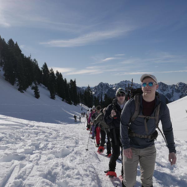 A group of PLU hikers in winter gear climbs a snowy mountain trail under a clear blue sky. They are surrounded by evergreen trees and distant peaks.