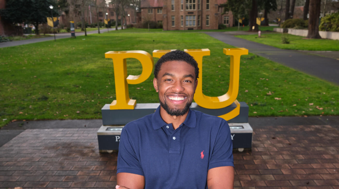 Andre Jones stands smiling while in front of the PLU sign on campus with is arms crossed.