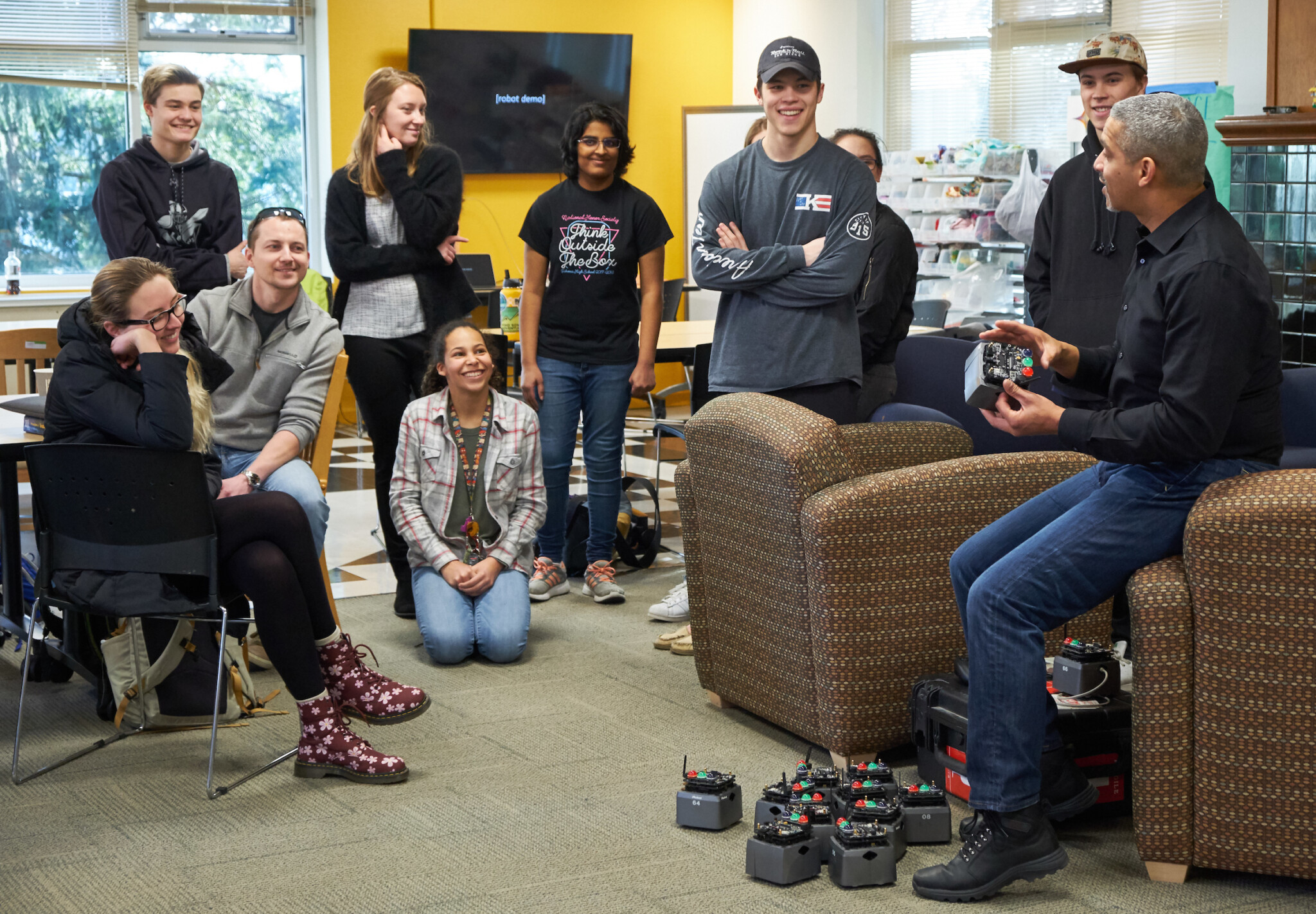 Students sit on the floor and stand in a room listening to a lecture from a professor.