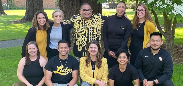 A group of PLU Admission Counselors wearing PLU gear pose together outside on campus