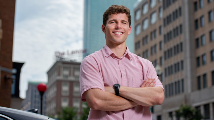 Keegan Dolan stands in Boston wearing a red shirt smiling at the camera. Behind him are buildings from Boston downtown.