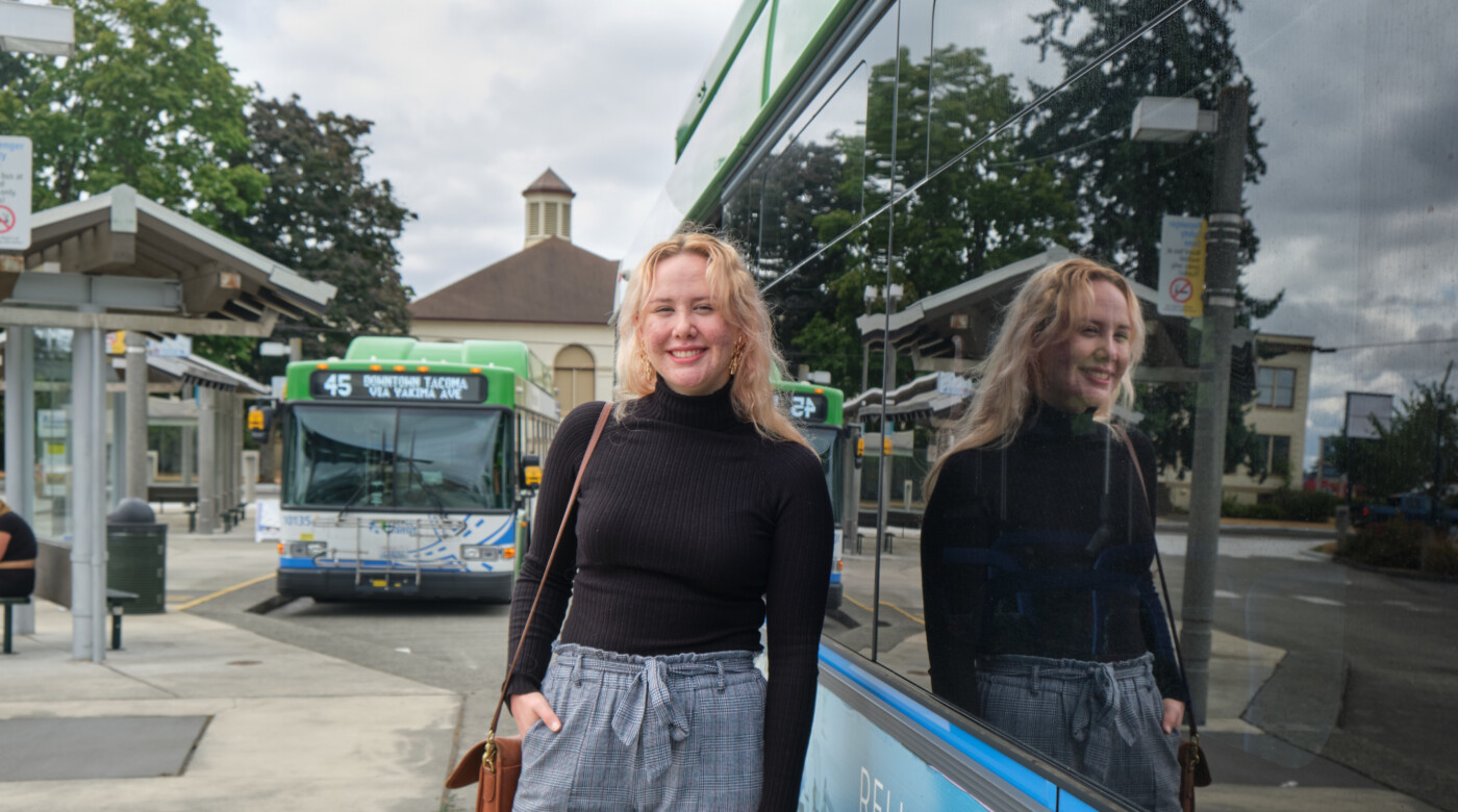 Student Kenzie Knapp stands in front of a city bus at the Tacoma Transit Center. Behind her is another city bus parked for passengers to board.