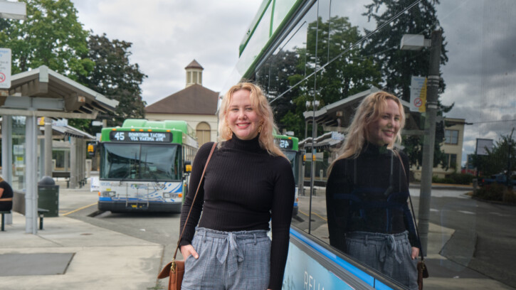 Student Kenzie Knapp stands in front of a city bus at the Tacoma Transit Center. Behind her is another city bus parked for passengers to board.