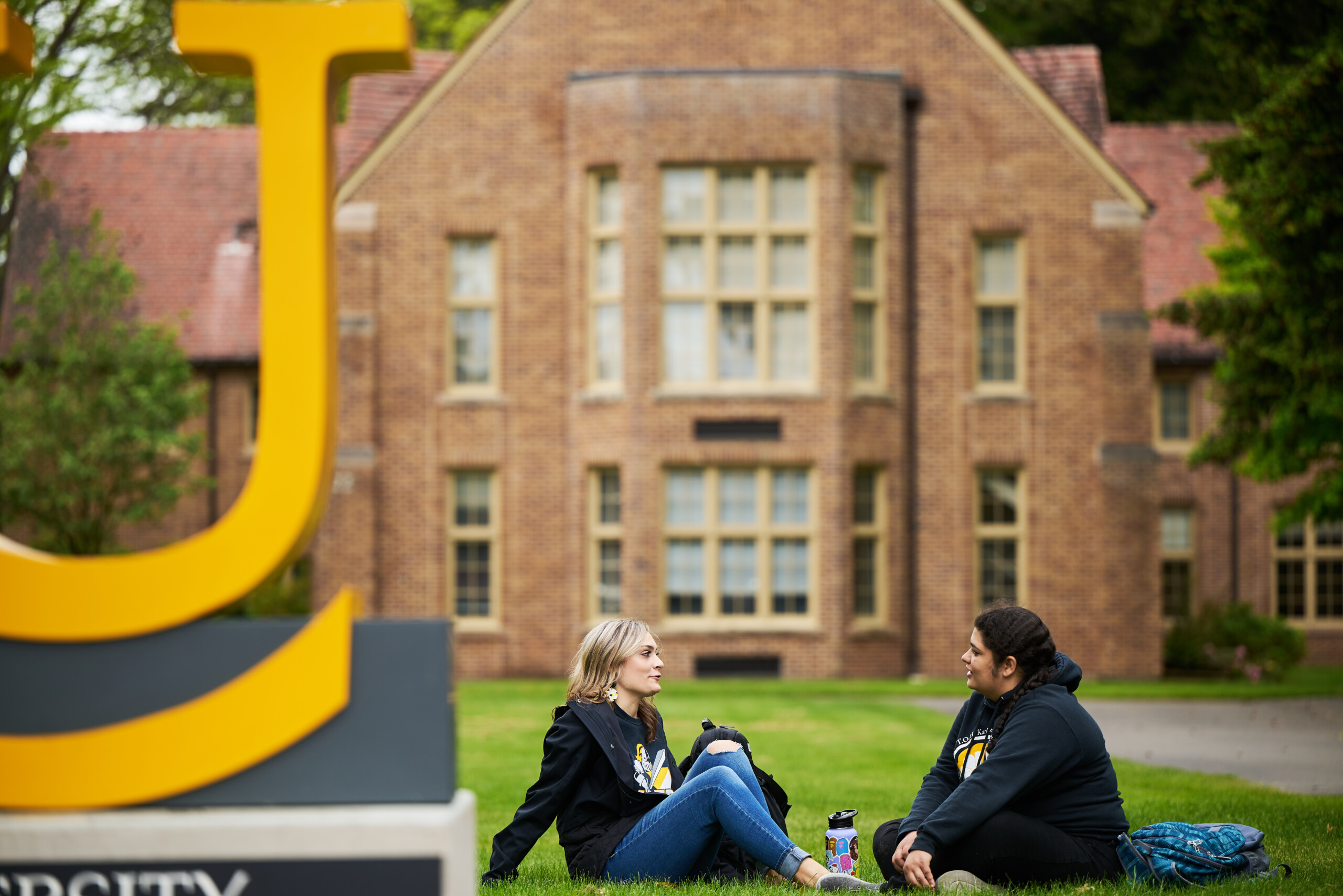 Two female students in black sweaters and jeans sit in the grass behind the PLU sign in front of a building.