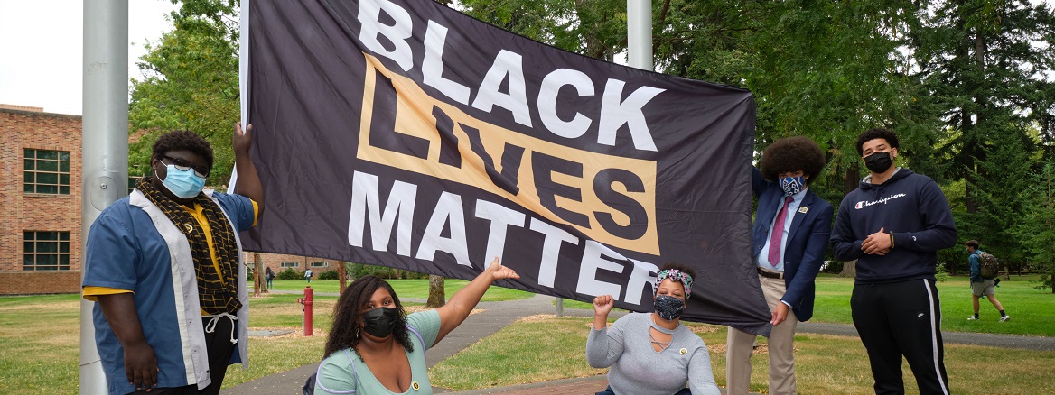 5 students wearing masks stand around a large 'Black Lives Matter' flag about to be raised on campus