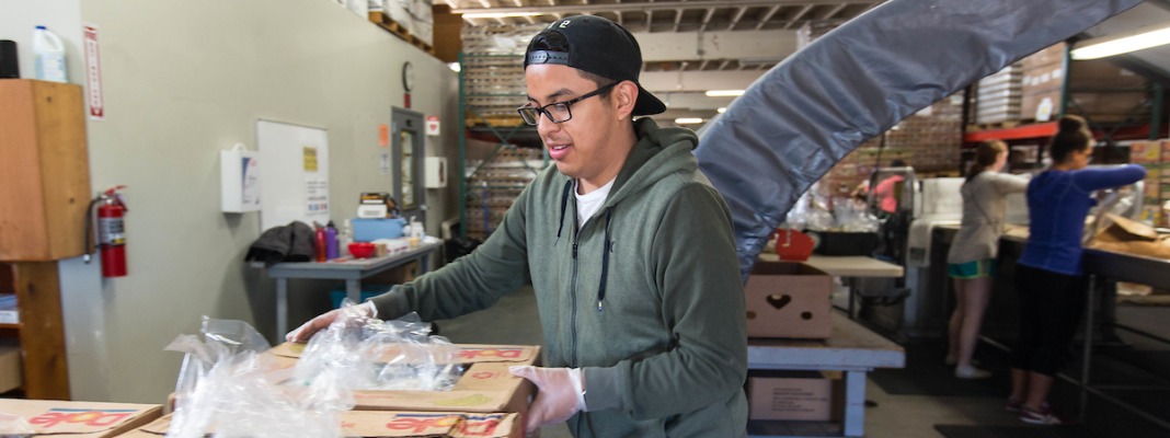 Student wearing glasses and backwards cap is stacking boxes inside a warehouse.