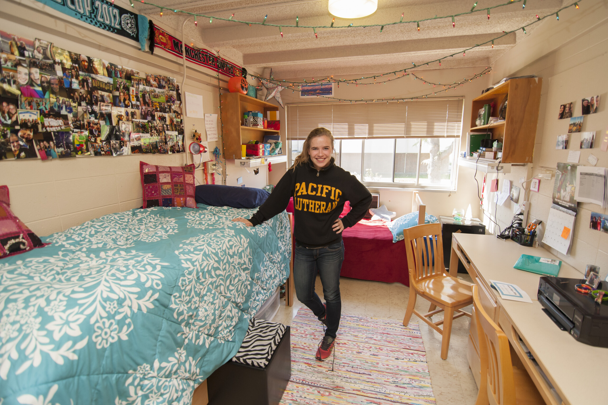 Female student stands in the middle of her dorm room wearing a black and yellow Pacific Lutheran University sweatshirts, blue jeans and red sneakers.