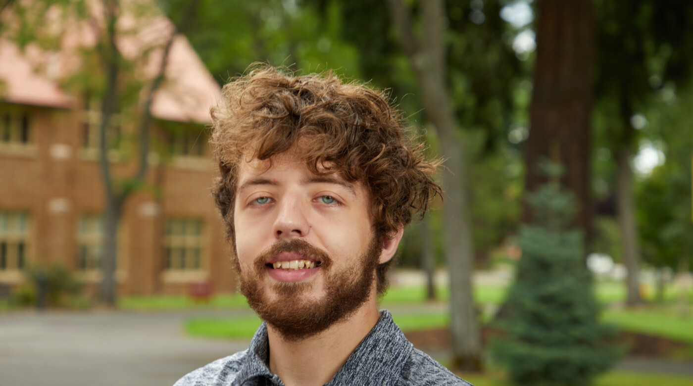 Ian Lindhartsen is seen standing on the PLU campus, looking at the camera wearing a black and white shirt.