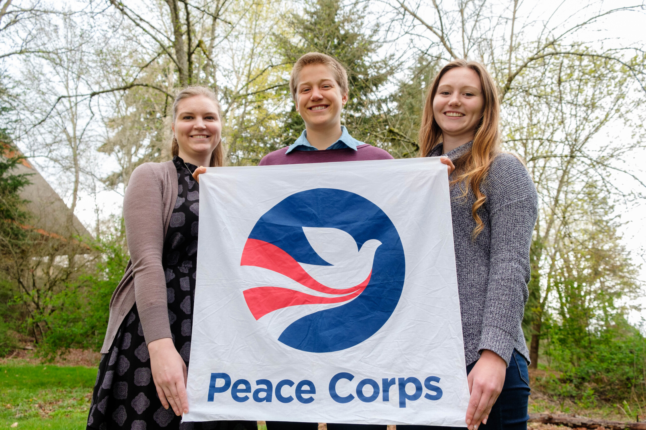Three students hold up a Peace Corps banner, with a the Peace corps logo, a dove on it.