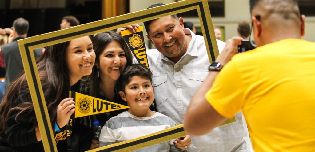 A mom, dad, daughter and young son smile and pose for a photo while holding a black and yellow frame and holding small PLU pennants. The photographer in a yellow shirt is also in the photo.