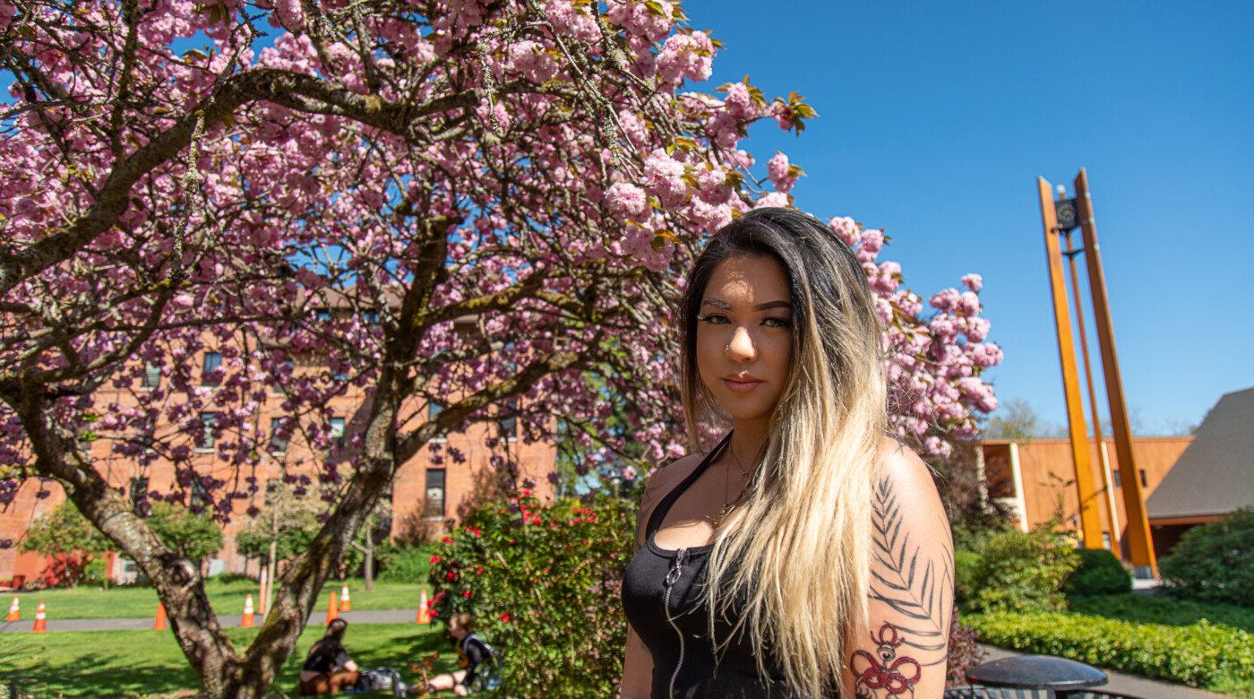 April Reyes poses at the camera while standing in front of a cherry tree on PLU campus next to the clock tower.
