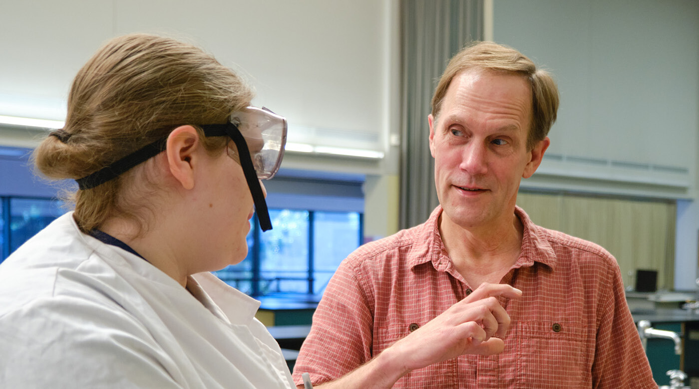 Professor Dean Waldo is speaking to an unidentified student wearing a lab coat and goggles.