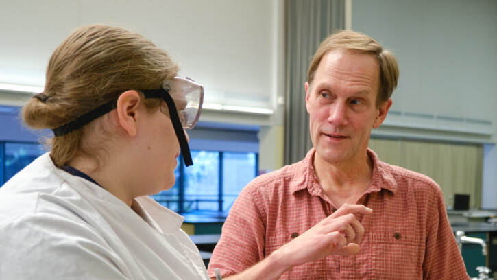 Professor Dean Waldo is speaking to an unidentified student wearing a lab coat and goggles.