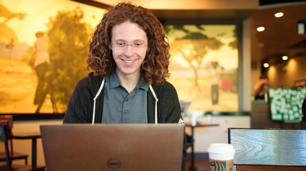 Matthew Conover smiles while sitting at his laptop in a coffee shop looking at the screen"
