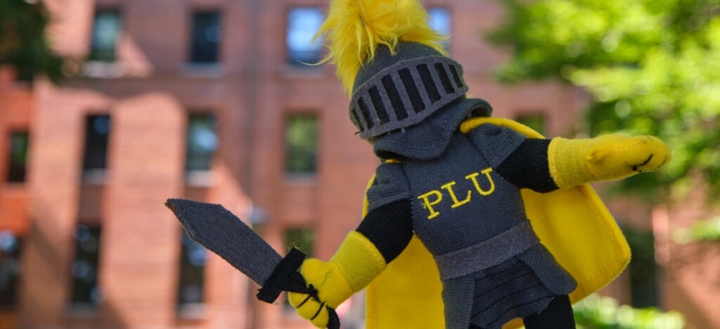 A close-up of a small stuffed PLU knight mascot outside. In the background is Harstad building.