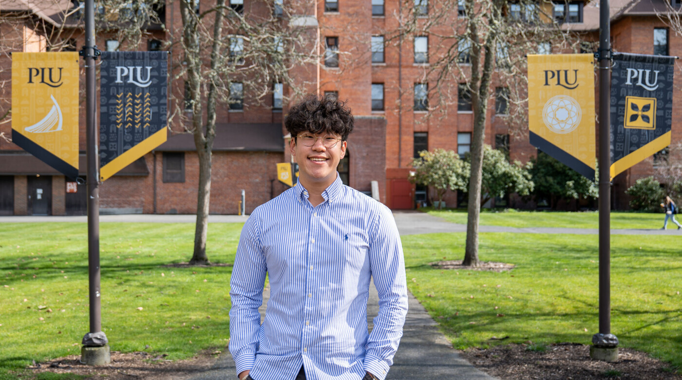 David Yun stands in the courtyard of the PLU campus wearing a blue shirt and glasses.