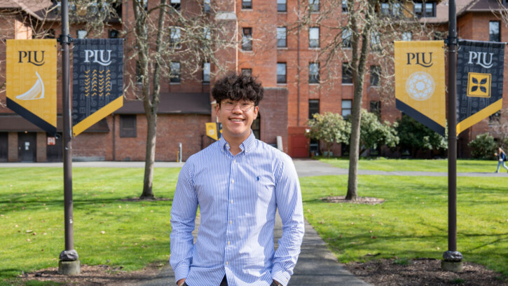 David Yun stands in the courtyard of the PLU campus wearing a blue shirt and glasses.