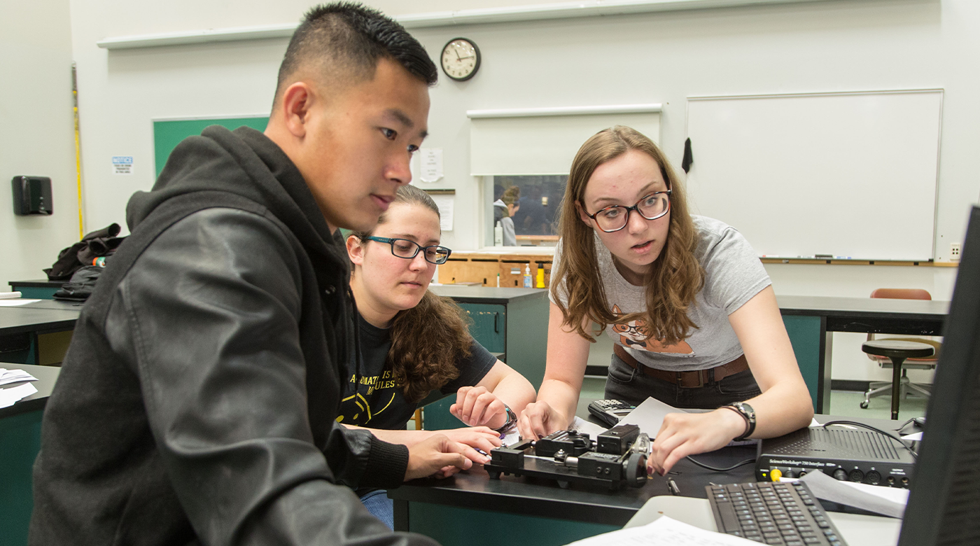 Three student, one male and two female, work on a robot while staring at a computer screen.