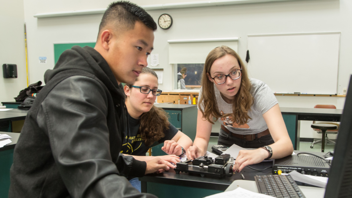Three student, one male and two female, work on a robot while staring at a computer screen.
