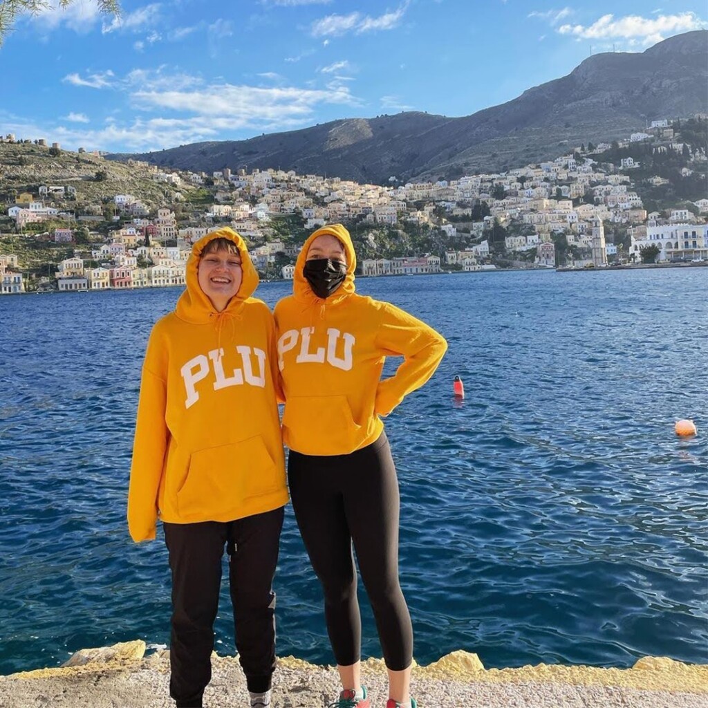 Two female students wearing yellow PLU hoodies stand in front of a body of water in Greece.