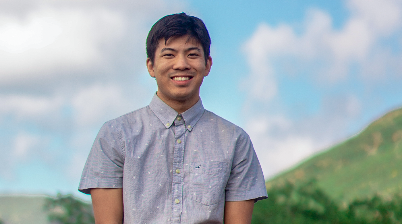 Cody Uehara smiles while standing in front of a stone fence overlooking a green valley.