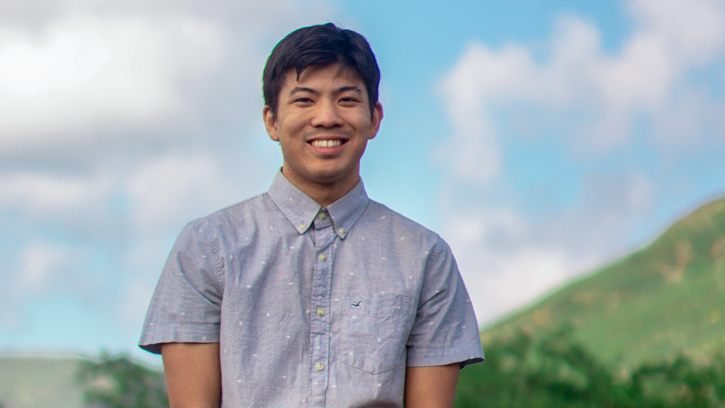 Cody Uehara smiles while standing in front of a stone fence overlooking a green valley.