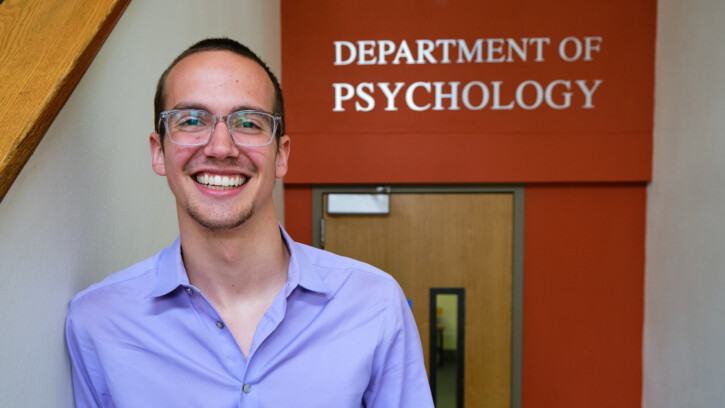 Nick Etzell stands in front of the Department of Psychology sign. Etzell is wearing a purple button shirt and glasses.