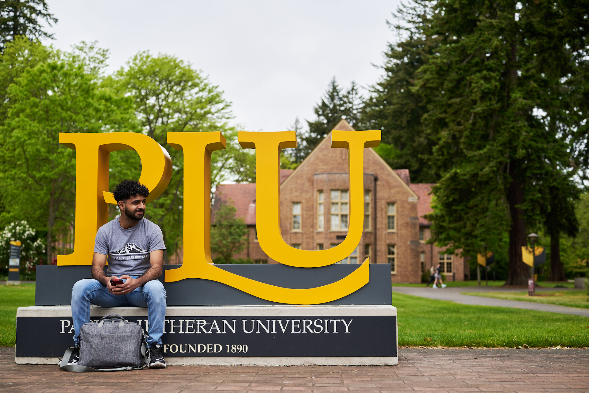 A student sits in front of a yellow PLU sign in front of a brick building surrounded by trees.