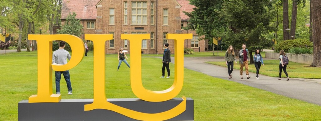 A large yellow PLU sign on campus, with students walking down the sidewalk beside it and other students playing frisbee on the grass behind the sign